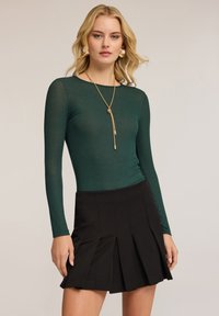 Tumma vihreä pitkähihainen paita, joka on valmistettu läpinäkyvästä materiaalista, yhdistetty mustaan laskoshameeseen. Kultaketju tuo yksityiskohtia asusteeseen.