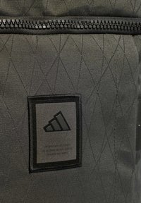 Musta tikattu kangas, jossa on geometrinen kuvio. Varusteena musta vetoketju ja suorakulmainen Adidas-logo-merkki, jossa on kolme raitaa.