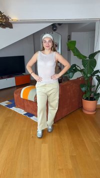 Νεαρή γυναίκα με κορδέλα στα μαλλιά, αμάνικο τοπ και μπεζ παντελόνι στέκεται χαμογελαστή σε ένα σαλόνι με ένα φυτό, καναπέ και τηλεόραση στο φόντο.