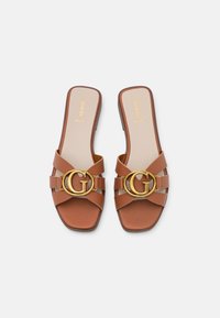 Braune Leder-Slidesandalen mit quadratischer Zehenpartie, ausgestattet mit goldenen, runden Logoakzenten und doppelten Kreuzriemen. Flache Sohle mit beiger Innensohle.