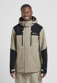 Giacca impermeabile beige e nera con colletto alto, cerniere frontali e cappuccio regolabile; presenta pannelli e loghi a contrasto.