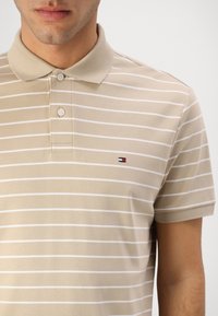 Beige polo-shirt met witte horizontale strepen, knoopsluiting en klein logo op de linkerborst. Gemaakt van zachte, geweven stof.