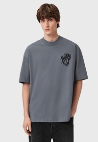 Camiseta de algodão cinza com decote redondo e mangas curtas. Apresenta um logótipo gráfico preto no lado esquerdo do peito. Ajuste descontraído e textura suave.