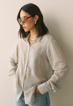 Moteris su tamsiais vidutinio ilgio plaukais, dėvinti didelius torto formos saulės akinius, auksinius lankelio formos auskarus, dryžuotą marškinių-kombinezoną ir šviesiai mėlynas džinsines kelnes.