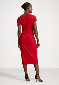 Rochie roșie, fără umeri, realizată dintr-un material elastic, cu detalii pliate pe laterale, asortată cu sandale negre cu toc înalt.