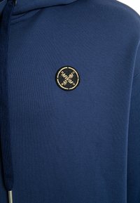Sudadera con capucha de color azul marino hecha de tela suave, con un logo bordado circular en acentos dorados en el pecho, y capucha con cordones.