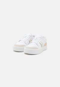 Fehér sneaker sima bőrből és bézs velúrból készült díszítésekkel. Vastag fehér talppal és színazonos fűzőkkel rendelkezik. Finom színblokk mintázat.