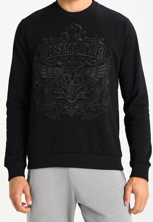 Sweat-shirt noir avec lion ailé en relief et texte « Versace Jeans », porté par une personne en pantalon gris clair.
