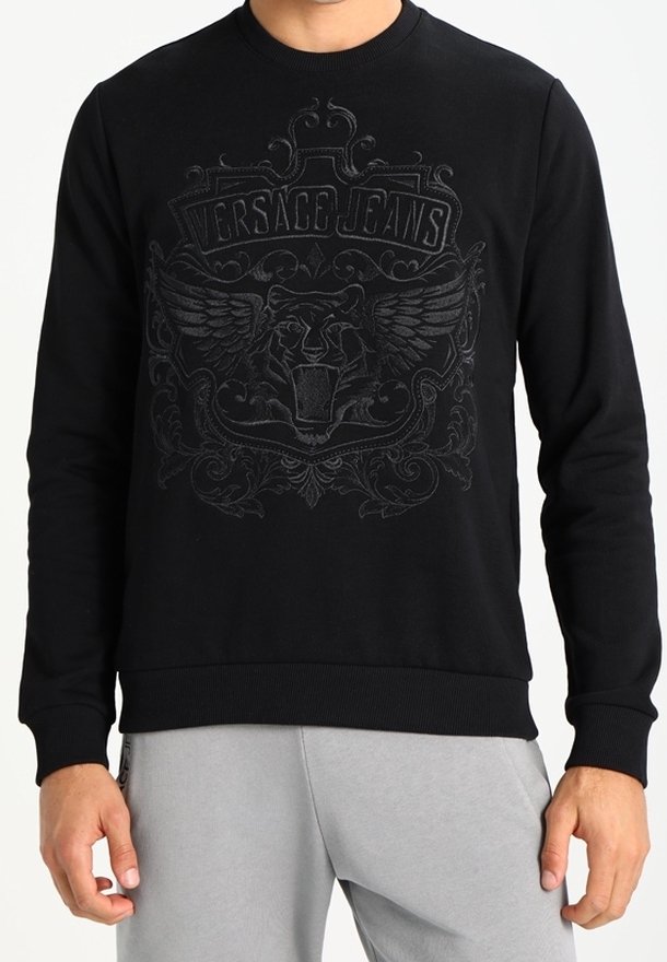 Sweat-shirt noir avec lion ailé en relief et texte « Versace Jeans », porté par une personne en pantalon gris clair.