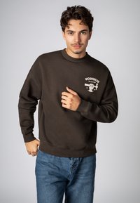 Brown sweatshirt with crew neckline, long sleeves, and ribbed cuffs. Front graphic reading "WORKWEAR COLLECTION." Worn with blue denim jeans.