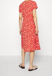 Vestido floral rojo con mangas cortas, corte a medio largo, que presenta un diseño fluido y sutiles patrones de flores blancas. Se lleva con sandalias planas.