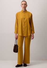 Set košile a kalhot v hořčičně žluté barvě, s límcem na knoflíky, dlouhými rukávy a volným střihem. Doplněno tmavě hnědou taškou.