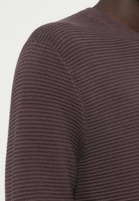 Κοντινό πλάνο στον ώμο και τον άνω βραχίονα ενός ατόμου με σκούρο δέρμα, που φοράει έναν καφέ πλεκτό πουλόβερ με ρίγες, σε ένα απλό ανοιχτό φόντο.