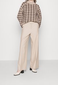 Beige Strickpullover mit braunem Hahnentrittmuster, kombiniert mit cremefarbenen weit geschnittenen Hosen und schwarzen Blockabsatzschuhen.