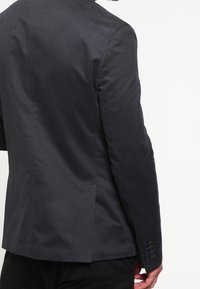 Mężczyzna w dopasowanej ciemnej marynarce, pokazujący szczegóły pleców i prawego ramienia z guzikami na rękawie na jednolitym białym tle.