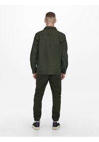 Olijfgroene jas met een gebuttonde kraag, gecombineerd met bijpassende joggers. De outfit heeft een ontspannen pasvorm en een gestructureerde stof. Zwarte en paarse sneakers.