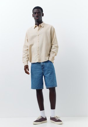 Junger Mann trägt beige langärmliges Hemd mit Knöpfen, blaue Jeansshorts, weiße Socken und dunkelviolette Sneakers, steht vor einem schlichten weißen Hintergrund.