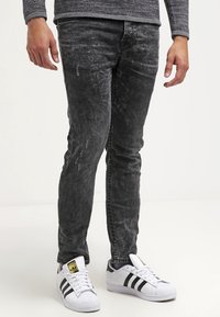 Jean en denim noir avec une texture délavée et un léger effet usé, associés à des baskets blanches avec des rayures noires et une semelle texturée.