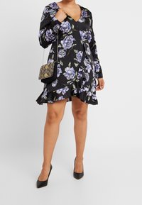 Svart blommig klänning med långa ärmar, V-ringning och volangkant. Lila blommor och gröna blad bildar ett djärvt mönster. Matchad med svarta klackar och en liten väska.
