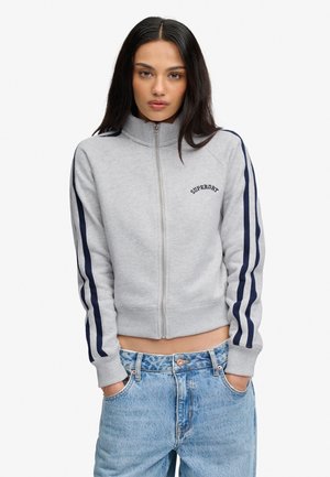 Giovane donna con lunghi capelli scuri che indossa un maglione grigio chiaro con cerniera e righe blu navy e jeans blu, mani in tasca, espressione neutra.