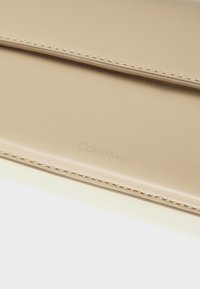 Calvin Klein beige skinnlommebok med jevn tekstur, subtil søm og preget logo. Enkel, rektangulær design.