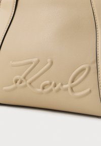 Seção de bolsa de couro bege com o logótipo em cursivo "Karl" em relevo e costura visível ao longo das bordas.