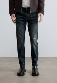 Sötétkék farmernadrág kopott részletekkel, fehér pólóval és fekete bőr csizmával viselve. Középmagas szabás, egyenes szárú kialakítás.