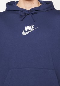 Mörkblå hoodie tillverkad av bomullsblandat tyg, med en framficka, dragsko på huva och silverfärgad Nike-logotyp. Slät textur, klassisk passform.