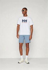 White T-shirt with bold "HH" and "Helly Hansen" logo in navy and orange, paired with light blue shorts and white sneakers.