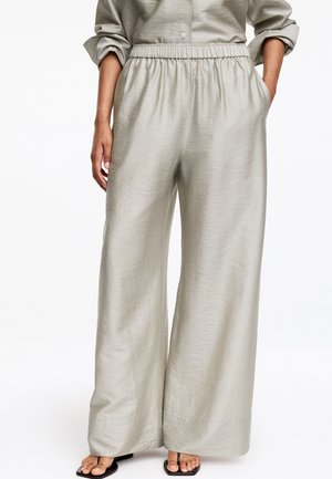 Pantalon gris clair à jambes larges avec taille élastique, porté avec des sandales noires à bout carré, mains dans les poches et chemise à manches longues retroussées.