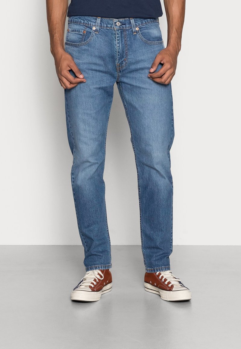 Jeans de mezclilla azul claro con un corte recto, textura deslavada, cinco bolsillos y herrajes metálicos. Llevando unos tenis marrones y blancos.