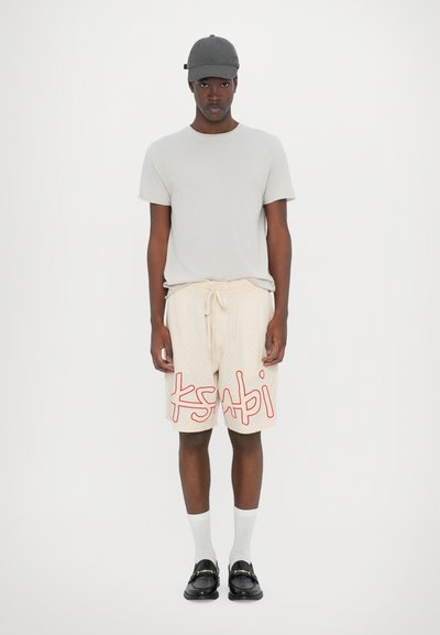 Camiseta gris claro, pantalones cortos crema con un logo rojo de "ksabi", calcetines blancos y zapatos negros. Los pantalones cortos presentan un diseño texturizado.