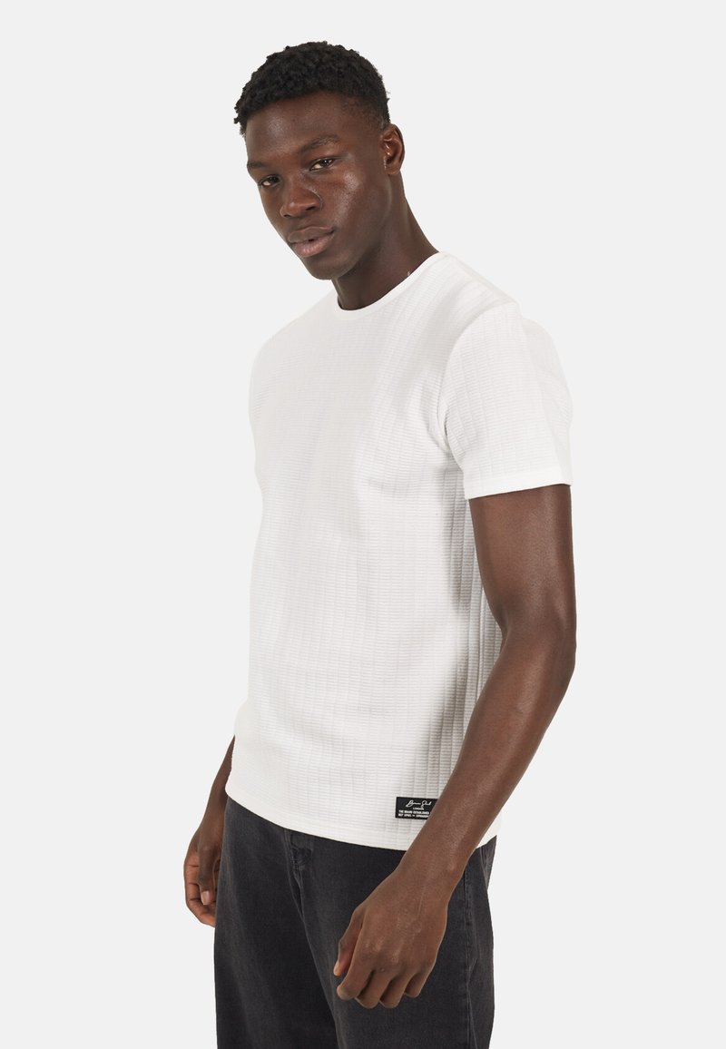 Mężczyzna w teksturowanej białej koszuli z krótkim rękawem i czarnych spodniach, stoi lekko obrócony na tle jasnego, jednolitego tła.