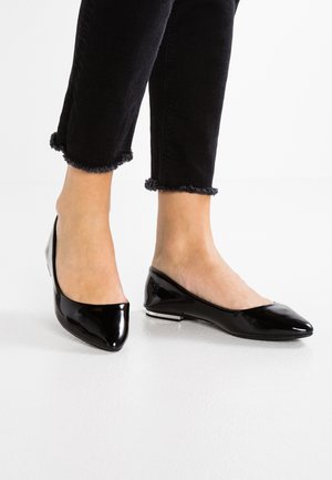 Person iført sorte korte bukser og skinnende sorte ballerinaer med spids tå, stående på en hvid overflade.