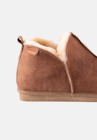 Chausson en suède marron avec une doublure en fourrure crème douce, une couture sur le côté et une étiquette embossée pour l'identification de la marque.
