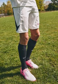Fehér Nike rövidnadrág fekete kiegészítőkkel, fekete térd fölé érő zoknival és rózsaszín focicipővel, zöld fűn állva.
