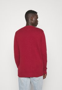 Maglione rosso lavorato a maglia con una finitura testurizzata, scollatura a girocollo e orli a coste, indossato con jeans azzurri chiari. La parte posteriore presenta un delicato dettaglio a V.