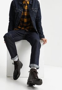 Una persona è seduta su un blocco bianco, indossando una giacca in denim scuro, una camicia a scacchi gialla e nera, jeans scuri arrotolati sui polsini, calzini grigi e stivali in pelle marrone scuro con lacci.