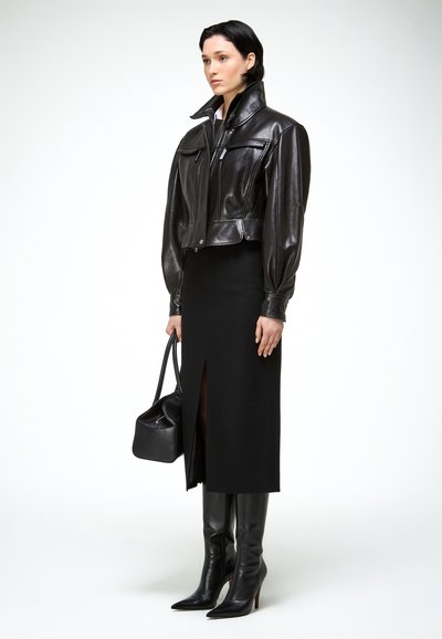 Žena v čiernej koženej bunde, čiernej midi sukni s predným rozparkom, čiernych čižmách po kolená a držiaca čiernu kabelku.