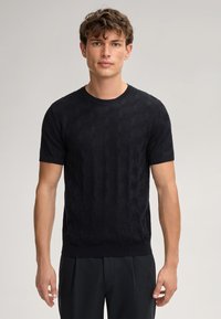 Maglione corto a maniche corte nero in maglia con un motivo geometrico testurizzato, scollatura a girocollo e orlo a coste. Indossato con pantaloni scuri.