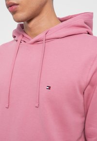 Różowa bawełniana bluza z kapturem ze sznurkiem oraz małym haftowanym logo w niebieskim, czerwonym i białym kolorze na lewej stronie klatki piersiowej.