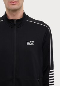 Giacca nera con zip realizzata in un tessuto morbido, con piping bianco sulle spalle e un piccolo logo EA7 sul petto.