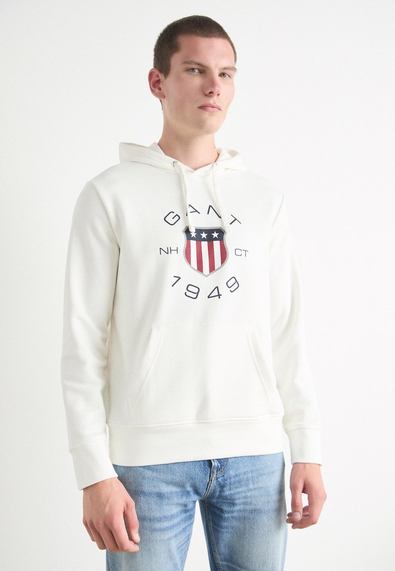 Kremfarget hettegenser med frontlomme, snorhette og et grafisk logo med designskjold i rødt, hvitt, blått og svart.
