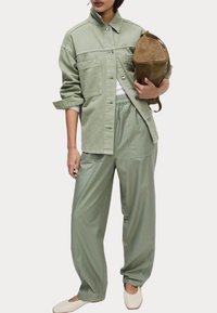 Lichtgroene oversized shirtjacket met twee voorzakken, gecombineerd met bijpassende loszittende broek. Een tan suede tas en crèmekleurige flats maken de look compleet.