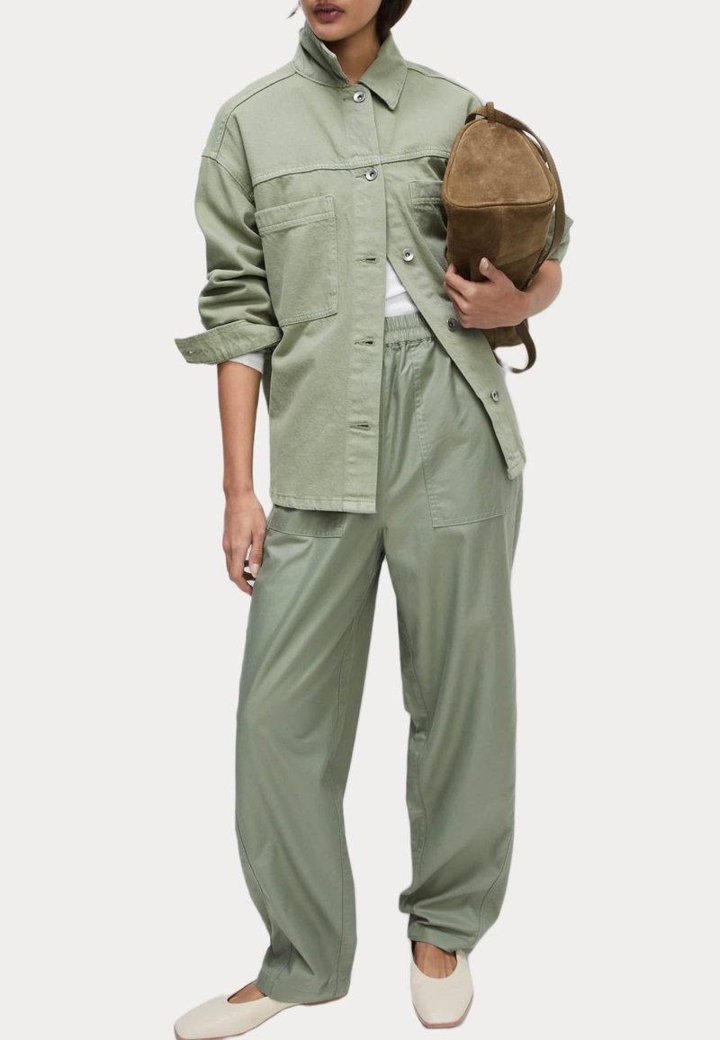 Lichtgroene oversized shirtjacket met twee voorzakken, gecombineerd met bijpassende loszittende broek. Een tan suede tas en crèmekleurige flats maken de look compleet.
