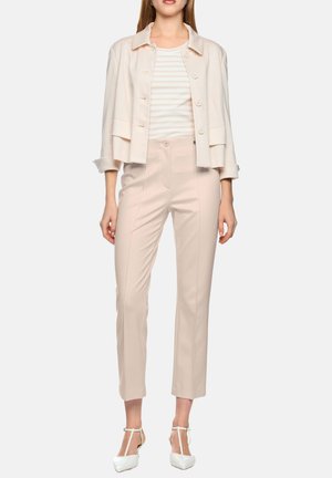 Frau trägt eine hellbeige, cropped Jacke, ein gestreiftes cremeweißes Top, beige Hosen mit hoher Taille und weiße T-Riemchen-Pumps.