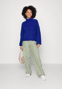 Μπλε πλεκτός πουλόβερ με ψηλό γιακά, παντελόνι με πράσινο και λευκό σχέδιο κυνόδοντα, λευκά αθλητικά παπούτσια και ανοιχτό ροζ τσάντα ώμου.