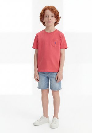 Rødhåret dreng står iført en koralfarvet kortærmet skjorte med lomme, lyse denimshorts og hvide sneakers mod en hvid baggrund.