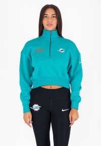 Felpa cropped color teal con colletto alto, caratterizzata dalla scritta "Miami Dolphins" ricamata in arancione e un logo sul davanti. Abbinata a leggings neri.