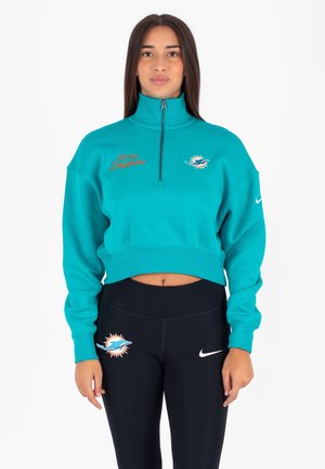 Felpa cropped color teal con colletto alto, caratterizzata dalla scritta "Miami Dolphins" ricamata in arancione e un logo sul davanti. Abbinata a leggings neri.